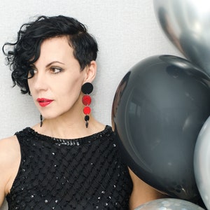 May include: A woman with short, curly black hair wearing a black sequined dress and red and black earrings. She is standing next to a group of balloons, including a black balloon, a silver balloon with confetti, and a silver balloon.