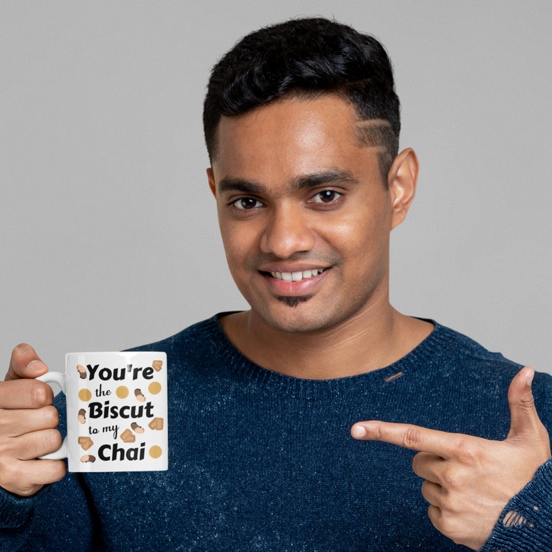 May include: White ceramic mug with the text "You're the Biscuit to my Chai" in black font. The mug features illustrations of biscuits and chai. The mug is held by a person wearing a blue sweater.