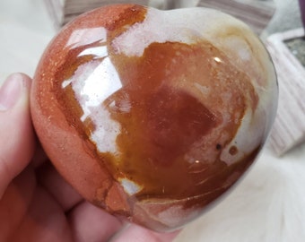 May include: A polished heart-shaped stone with a mix of red, brown, and white hues. The stone has a glossy finish, reflecting light. The heart is held in a hand, with small potted plants in the background. A decorative item.