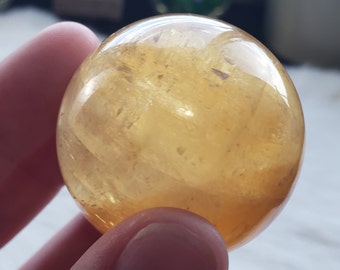 May include: A polished, translucent citrine crystal sphere. The sphere is a warm, golden-yellow color with internal patterns and inclusions. The surface is smooth and reflective, catching the light.