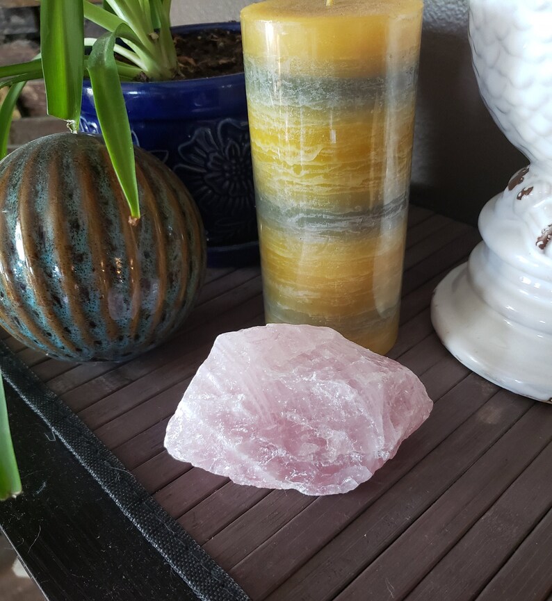 May include: A large, rough-cut rose quartz crystal sits on a wooden surface. A tall, multi-toned yellow and gray candle is behind the crystal. A blue ceramic pot and a decorative white owl figurine are also in the background.