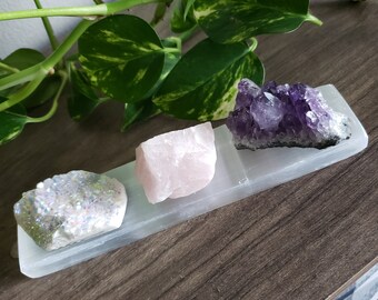 May include: A selenite crystal charging plate holds three raw crystals: a shimmering white druzy, a light pink rose quartz, and a deep purple amethyst. The crystals are arranged on a rectangular white plate, with green leafy plants in the background.