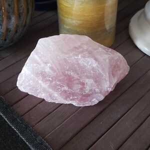 May include: A large, rough-hewn chunk of rose quartz, displaying a soft pink hue. The stone has a crystalline structure and sits on a dark brown surface. Other objects are visible in the background.