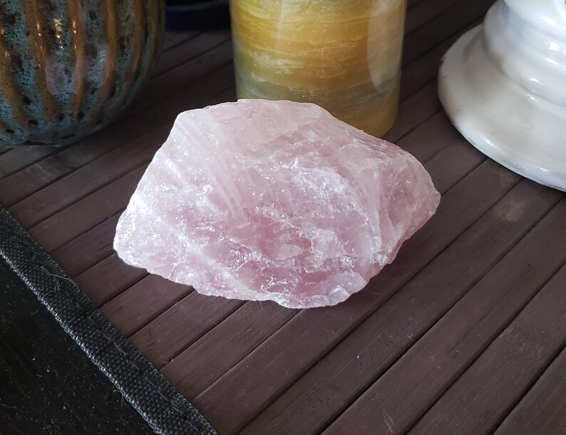 May include: A large, rough-hewn chunk of rose quartz, displaying a soft pink hue. The stone has a crystalline structure and sits on a dark brown surface. Other objects are visible in the background.