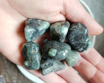 May include: A collection of tumbled emerald stones, showcasing a range of dark green hues with some areas of black and gray. The stones have a smooth, polished appearance, and are held in a hand.