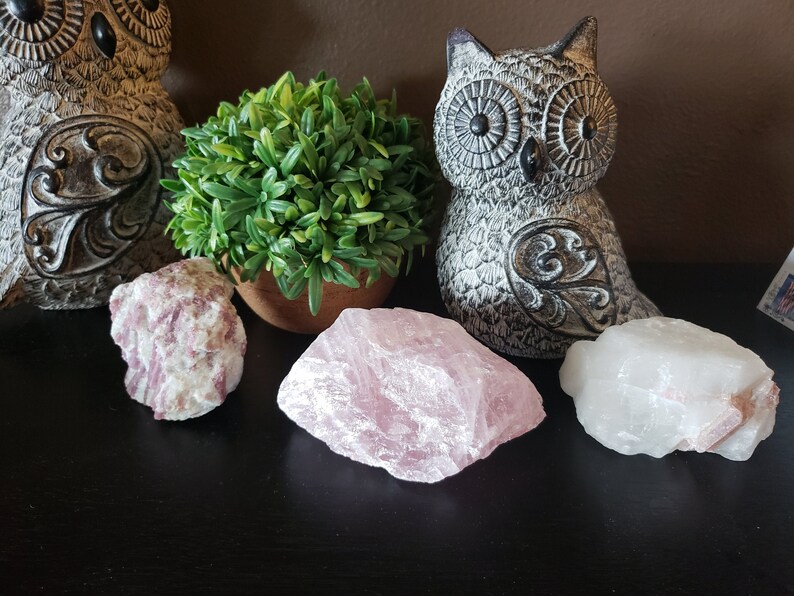 May include: A collection of crystals and decorative owl figurines. The crystals include a large, light pink rose quartz, a white crystal with pink inclusions, and a smaller pink and white crystal. A small potted plant sits between the crystals.