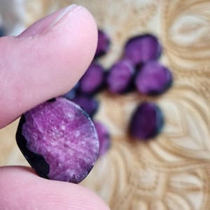 May include: A close-up of a faceted, oval gemstone held between fingers. The gem is a deep magenta colour with black edges and a textured surface. Other similar gems are blurred in the background.