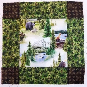 May include: A patchwork quilt block with a green and brown floral print. The centre of the block features a white background with a mountain scene, a bear, and a seal.
