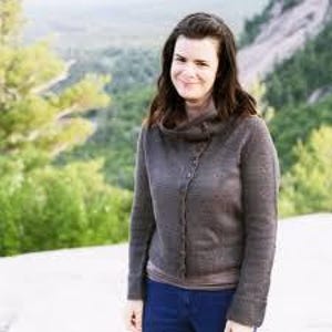 May include: A woman wearing a dark gray knitted cardigan with a cowl neck and button closure. The cardigan is paired with a light brown top and blue jeans. The background features a blurred view of trees and a mountain.