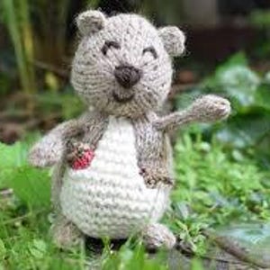 May include: A handmade knitted bear toy with a light brown body, a white belly, and a small red strawberry. The bear has a smiling face and is waving its right arm. The toy is set against a backdrop of green foliage.