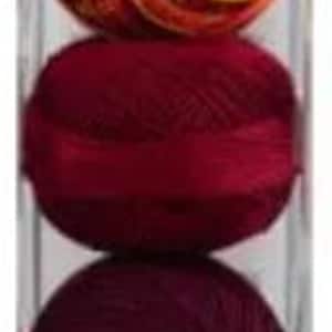 Könnte beinhalten: Eine transparente Kunststoffverpackung mit fünf Spulen Häkelgarn von Lizbeth in Orange-, Rot- und Brauntönen. Die Spulen sind eng gewickelt und vertikal gestapelt, wodurch eine Reihe warmer, erdiger Farbtöne präsentiert wird.