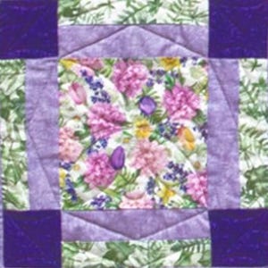 May include: A square quilt block featuring a central floral print with pink and purple flowers. The block is framed by lavender and purple fabric, with dark purple squares at each corner. The design includes green and white patterned borders.