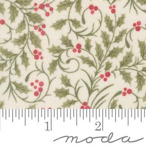 May include: A cream-colored fabric featuring a repeating pattern of green holly leaves and red berries. A white ruler is positioned at the bottom, showing a scale of two inches, with the word moda printed in a cursive script below the ruler.