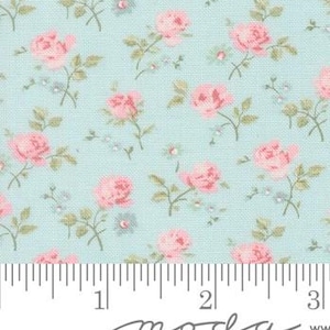 Op de afbeelding: Een close-up van een stof met een herhalend bloemenpatroon. Roze rozen en kleine blauwe bloemen zijn verspreid over een lichtblauwe achtergrond. De stof heeft een liniaal langs de onderkant, met het woord "moda" in cursief gedrukt.