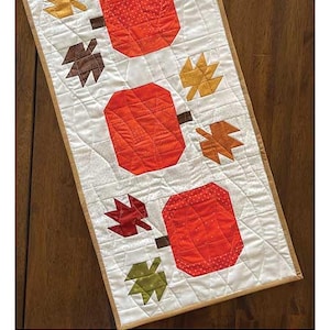 May include: A white quilted table runner with a brown border featuring a pattern of orange pumpkins and brown and yellow leaves. The text "Pumpkin Harvest Table Runner" is at the top of the runner. The finished size is 13 inches by 42 inches.