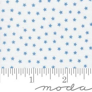 May include: White fabric with a repeating pattern of small, blue five-pointed stars. A ruler is visible along the bottom edge, marked in inches. The fabric is likely cotton or a similar material, suitable for quilting or crafting projects.