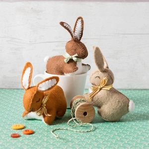 May include: Three felt bunny rabbit toys with white accents and yellow ribbon bows. The bunnies are brown, tan, and orange. The bunnies are sitting on a white cup and a green surface.