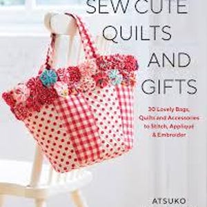 Peut inclure: Un sac fourre-tout fait à la main avec un motif vichy rouge et blanc et un motif à pois rouges. Le sac est décoré de fleurs en tissu rouges et roses et d'accents bleus. Le livre "SEW CUTE QUILTS AND GIFTS" est visible.