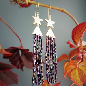 Könnte beinhalten: Goldene Stern-Ohrringe mit einem langen, baumelnden Fransen aus schimmernden violetten und schwarzen Perlen.