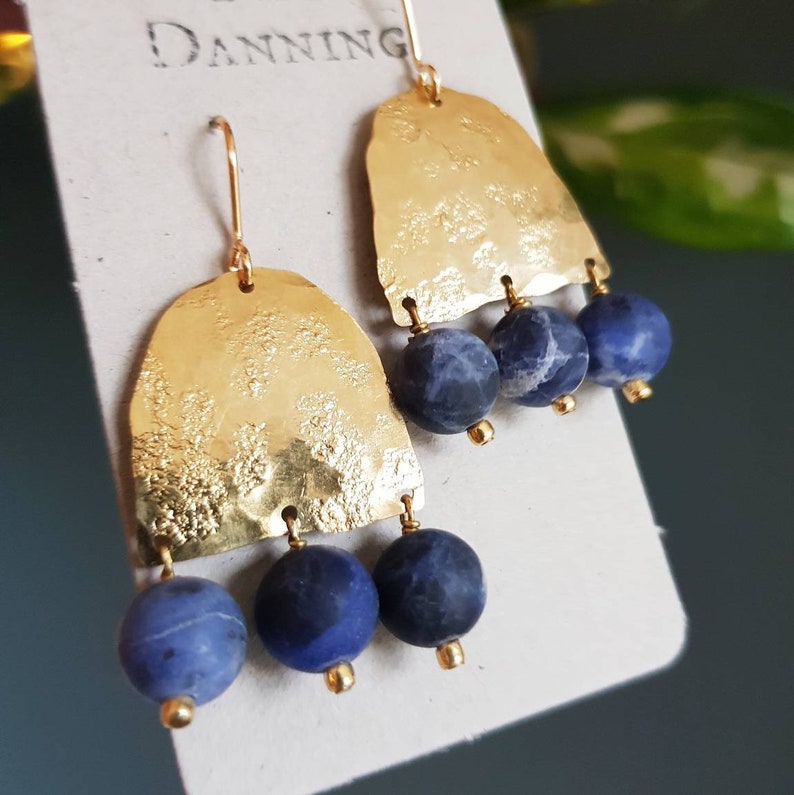 May include: A pair of gold-colored earrings with a textured, arch-shaped top and three dark blue, spherical beads hanging below. The earrings have a hook closure. The brand name "Danning" is printed on the product tag.