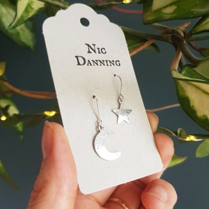 May include: A pair of silver earrings on a white card with the text "Nic Danning". One earring is a crescent moon and the other is a star.