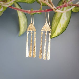 Könnte beinhalten: Goldene Dreiecks-Ohrringe mit drei dünnen, strukturierten, goldenen Stäben, die an jedem Dreieck hängen.