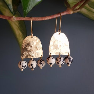 May include: Gold-tone earrings with a hammered texture and a curved top. Each earring features three round, speckled beads in black and white, suspended below the gold shape. The earrings hang from gold-colored ear wires.