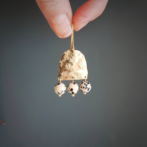 May include: Gold-toned earrings with a textured, bell-shaped top and three dangling, round beads. The beads are white with black spots. The earrings are held up by a gold hook.