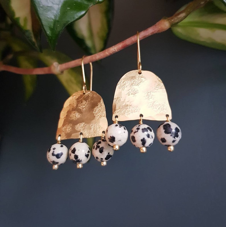May include: Gold-tone earrings with a hammered texture and a dangling design. Each earring features a gold arch shape and three Dalmatian jasper beads with black spots. The earrings hang from gold-colored ear wires.