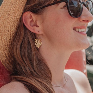 May include: A woman wearing a straw hat and sunglasses with gold leaf earrings shaped like a monstera leaf.