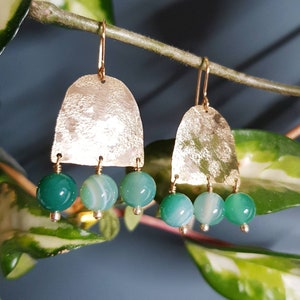 May include: Gold-coloured earrings featuring a textured, arched top and three hanging green and white agate beads. The earrings have a simple hook closure. The background is a blurred image of green leaves and a blue-grey wall.