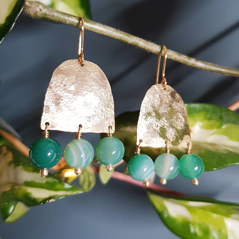 May include: Gold-coloured earrings featuring a textured, arched top and three hanging green and white agate beads. The earrings have a simple hook closure. The background is a blurred image of green leaves and a blue-grey wall.