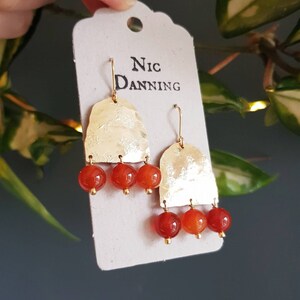May include: Gold-tone earrings with a hammered texture and three dangling orange-red beads. The earrings are on a white tag with the name "Nic Danning" printed in black. The earrings have a hook closure.