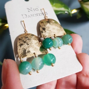 May include: Gold-coloured earrings with a hammered finish, each with three green and white striped beads. The earrings hang from a gold hook. The brand name "Nic Danning" is printed on the tag.