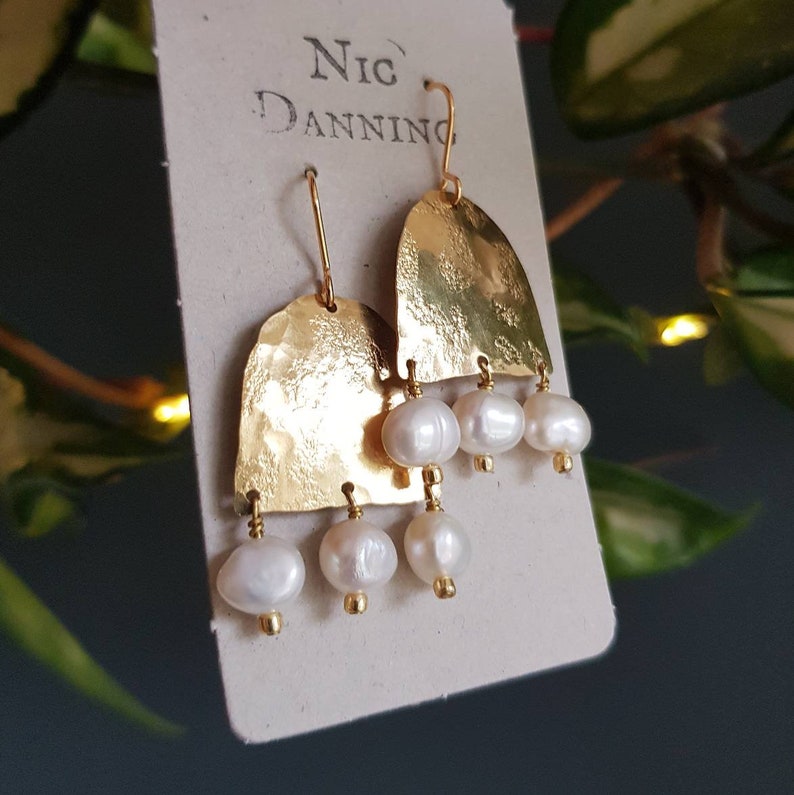 May include: Gold-toned, hammered metal earrings with a half-moon shape, each adorned with three white, pear-shaped pearls. The earrings hang from gold-colored hooks. The brand name "Nic Danning" is printed on the backing card.