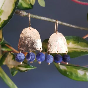 May include: Gold-tone earrings with a hammered texture and three dangling blue beads. The earrings hang from a gold hook. The background is a blurred image of green leaves and a blue wall.