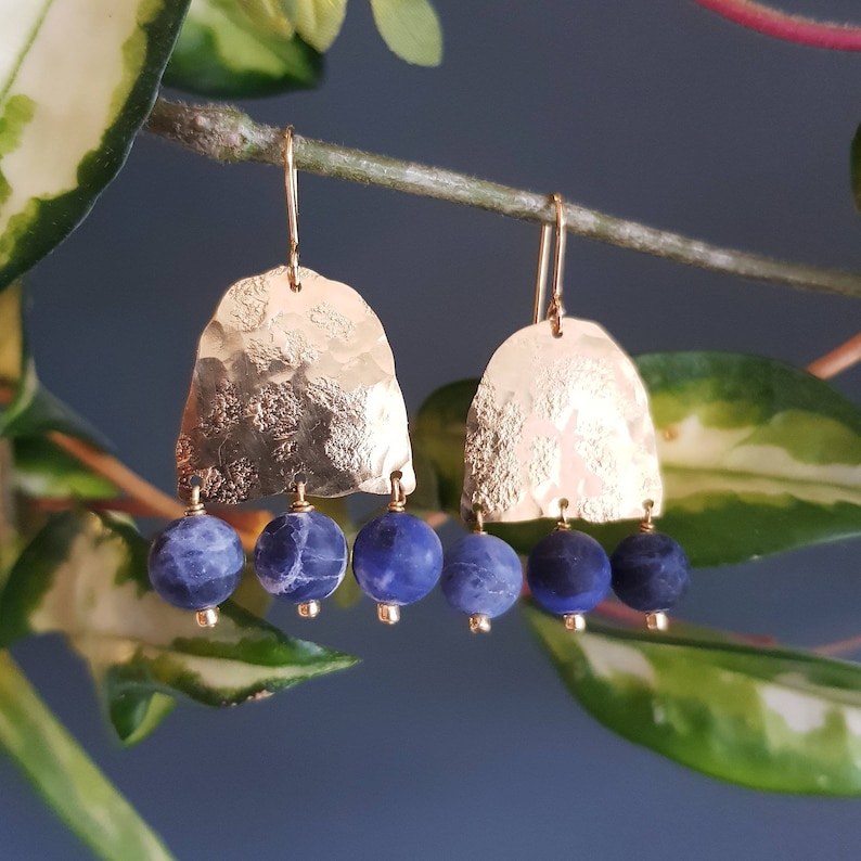 May include: Gold-tone earrings with a hammered texture and three dangling blue beads. The earrings hang from a gold hook. The background is a blurred image of green leaves and a blue wall.