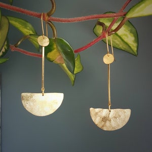 May include: A pair of gold-colored earrings with a unique design. Each earring features a small, textured disc at the top, connected to a long, thin bar. A textured, half-moon shape hangs from the bottom of the bar. The earrings are displayed against a backdrop of green leaves.