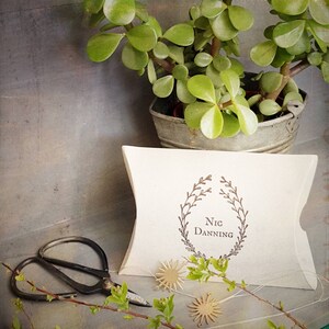 May include: A white pillow box with a brown wreath and the text "Nic Danning" printed on the front. The box is sitting on a table with a potted plant and a pair of scissors.
