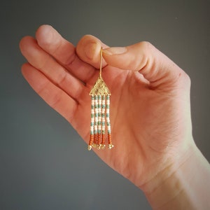 May include: Gold triangle earring with a beaded tassel. The tassel is made of white, turquoise, and orange beads.