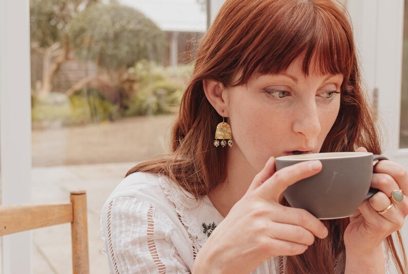 May include: A woman with red hair wearing gold earrings and rings, sipping from a gray mug. The earrings have a textured, bell-like shape with dangling beads. The woman is wearing a white blouse with embroidered details.