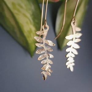 Puede incluir: Un par de pendientes dorados con forma de hoja. Los pendientes cuelgan de delicados alambres dorados. Las hojas tienen una superficie texturizada y se destacan sobre un fondo borroso verde y azul.
