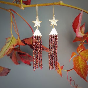 Könnte beinhalten: Goldene Stern-Ohrringe mit einem burgunderfarbenen Perlen-Quaste. Die Ohrringe hängen an einem Zweig mit Herbstblättern.