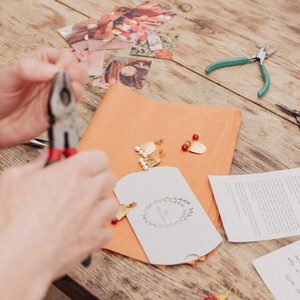 Pu&ograve; includere: Un tavolo di legno mostra materiali per la creazione di gioielli. Sono visibili pinze, forbici e un piccolo biglietto bianco con un motivo floreale. Orecchini dorati con perline rosse e bianche sono disposti su carta arancione.