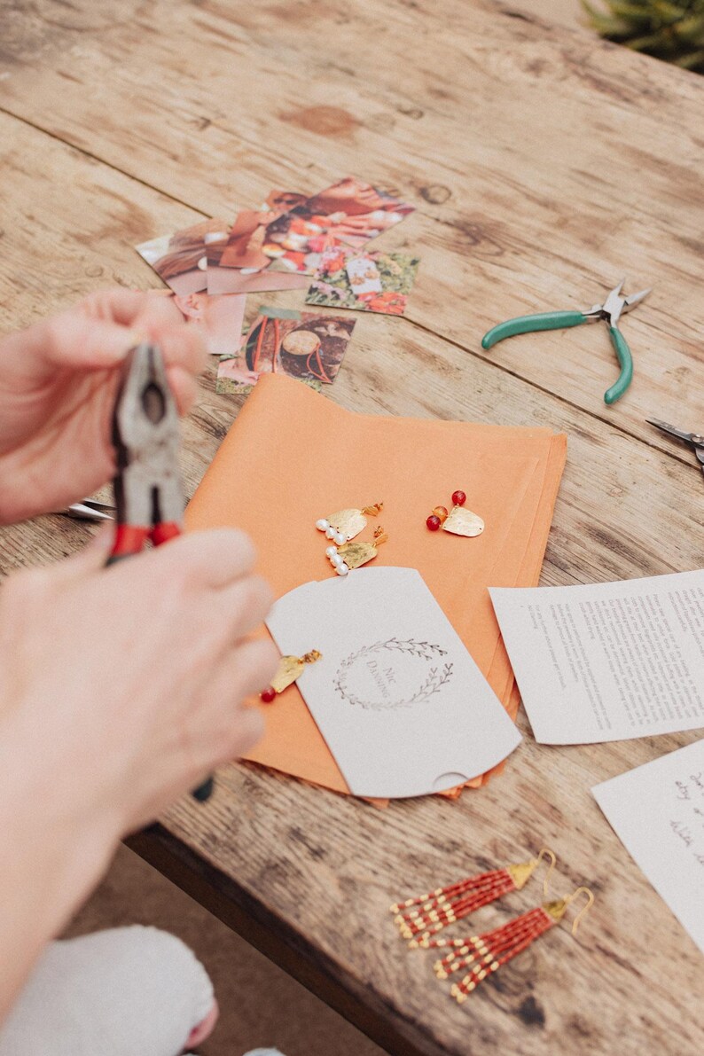 Pu&ograve; includere: Un tavolo di legno mostra materiali per la creazione di gioielli. Sono visibili pinze, forbici e un piccolo biglietto bianco con un motivo floreale. Orecchini dorati con perline rosse e bianche sono disposti su carta arancione.