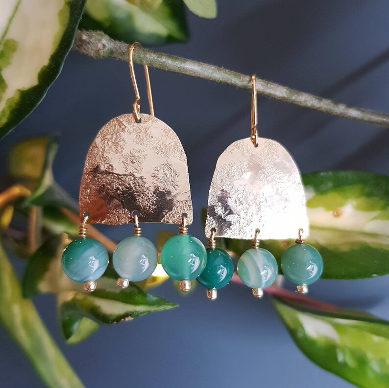 May include: Gold-coloured earrings featuring a textured, arched top and three hanging green and blue agate beads. The earrings have a simple hook closure. The background shows green leaves and a blue backdrop.
