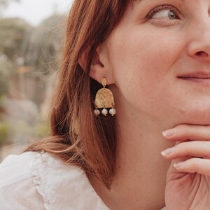 May include: Gold-toned earrings with a textured, arched design and three dangling, small, pearl-like beads. The earrings are worn by a person with red hair and a white shirt. A gold ring with a light blue stone is also visible.