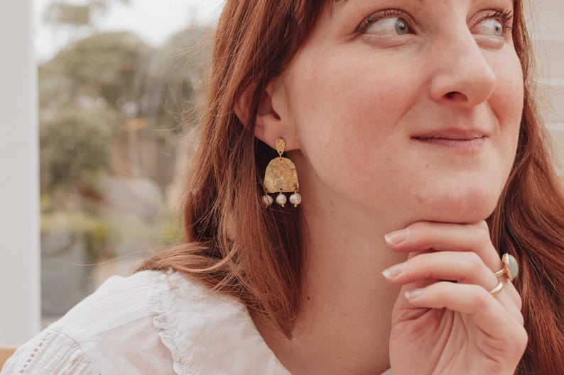May include: Gold-toned earrings with a textured, arched design and three dangling, small, pearl-like beads. The earrings are worn by a person with red hair and a white shirt. A gold ring with a light blue stone is also visible.