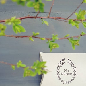 May include: A white, pillow-shaped box with the text "Nic Danning" inside a laurel wreath design. The box is set against a gray background, with a blurred branch of green leaves and brown stems in the foreground.