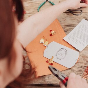 May include: Handmade gold earrings with white pearls and red beads are arranged on an orange surface. A small white box with a floral design and the text "Nic Darling" is also present. Pliers and other tools are visible, suggesting a craft setting.